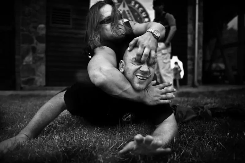 Jarosław Bielan przytrzymuje drugiego mężczyznę na ziemi, zakładając chwyt na głowie. Działanie odbywa się na trawniku przed budynkiem z drewnianą elewacją.