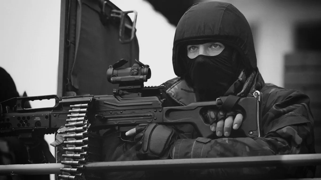 Osoba w czarnej odzieży taktycznej trzyma karabin maszynowy, skupiona na obserwacji otoczenia. Znajduje się w osłoniętym miejscu, gotowa do działania. Czarnobiała fotografia podkreśla napięcie sytuacji.