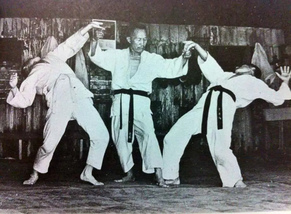 Trzech mężczyzn w strojach do sztuk walki, dwóch z nich wykonuje dramatyczne, wygięte ruchy podczas wspólnej techniki, trzeci stoi pośrodku. W tle drewniane ściany z wiszącymi ubraniami.