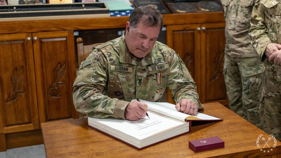 Żołnierz w mundurze z naszywką "Dzieran" pisze w dużej księdze na drewnianym biurku. W tle widoczne są drewniane szafki.