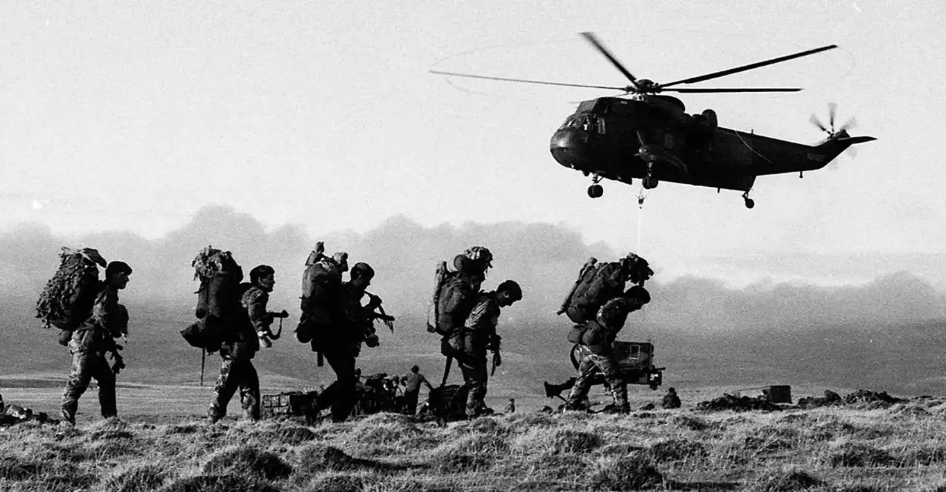 Żołnierze z dużymi plecakami idą w szyku bojowym. Nad nimi unosi się helikopter. Wszystko dzieje się na otwartej, trawiastej przestrzeni wśród gór.