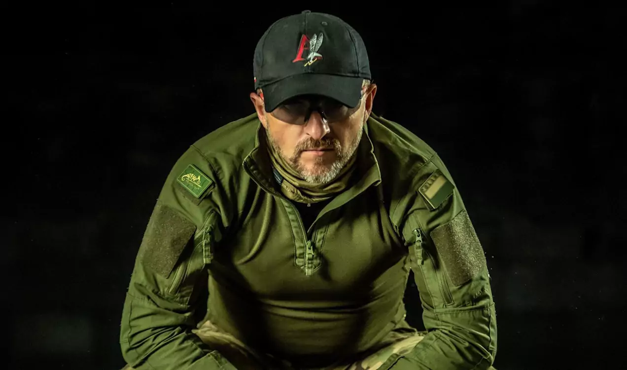 Mężczyzna w zielonej kurtce i bejsbolówce z wypukłym godłem ptaka patrzy w dół w skupieniu, siedząc w ciemnym otoczeniu. Na ramieniu widoczna naszywka z napisem "Wilk".