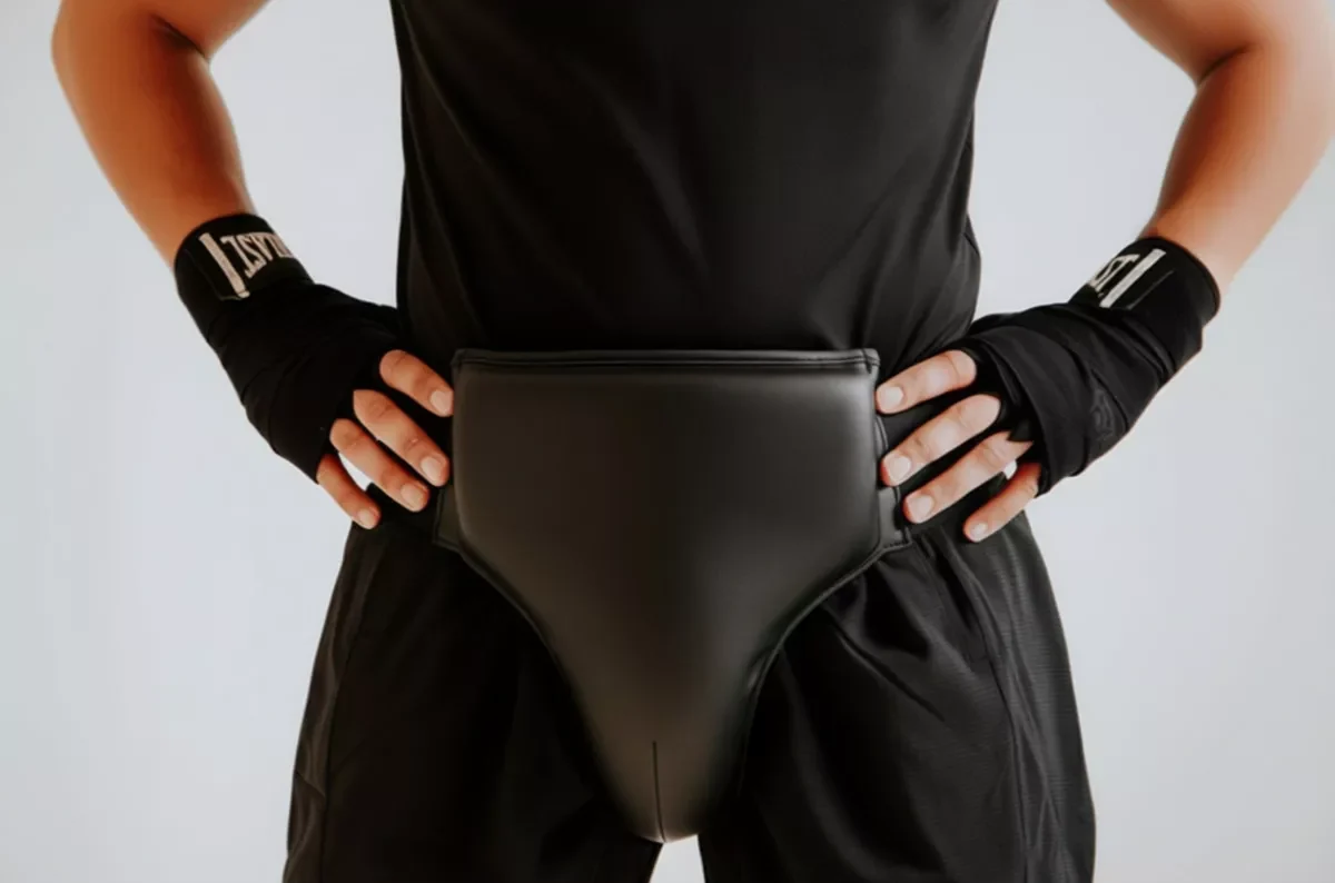 Osoba w czarnym stroju sportowym i ochraniaczu na krocze stoi w neutralnej pozie, z rękami na biodrach. Na nadgarstkach opaski z napisem "LONSDALE". Tło jasne i jednolite.