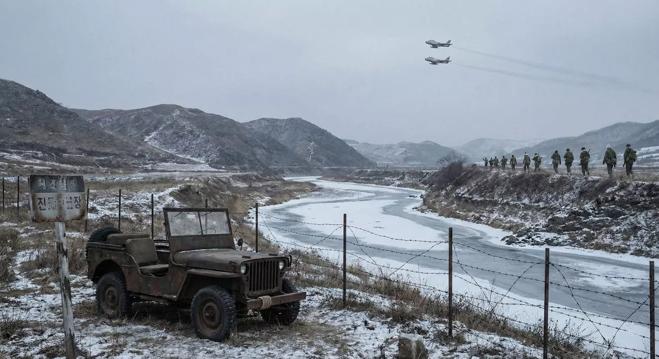 Jeep stoi nieruchomo w zimowym, górzystym krajobrazie obok zamarzniętej rzeki. Na niebie lecą dwa samoloty, a w oddali maszerują żołnierze. Tablica z tekstem po koreańsku.
