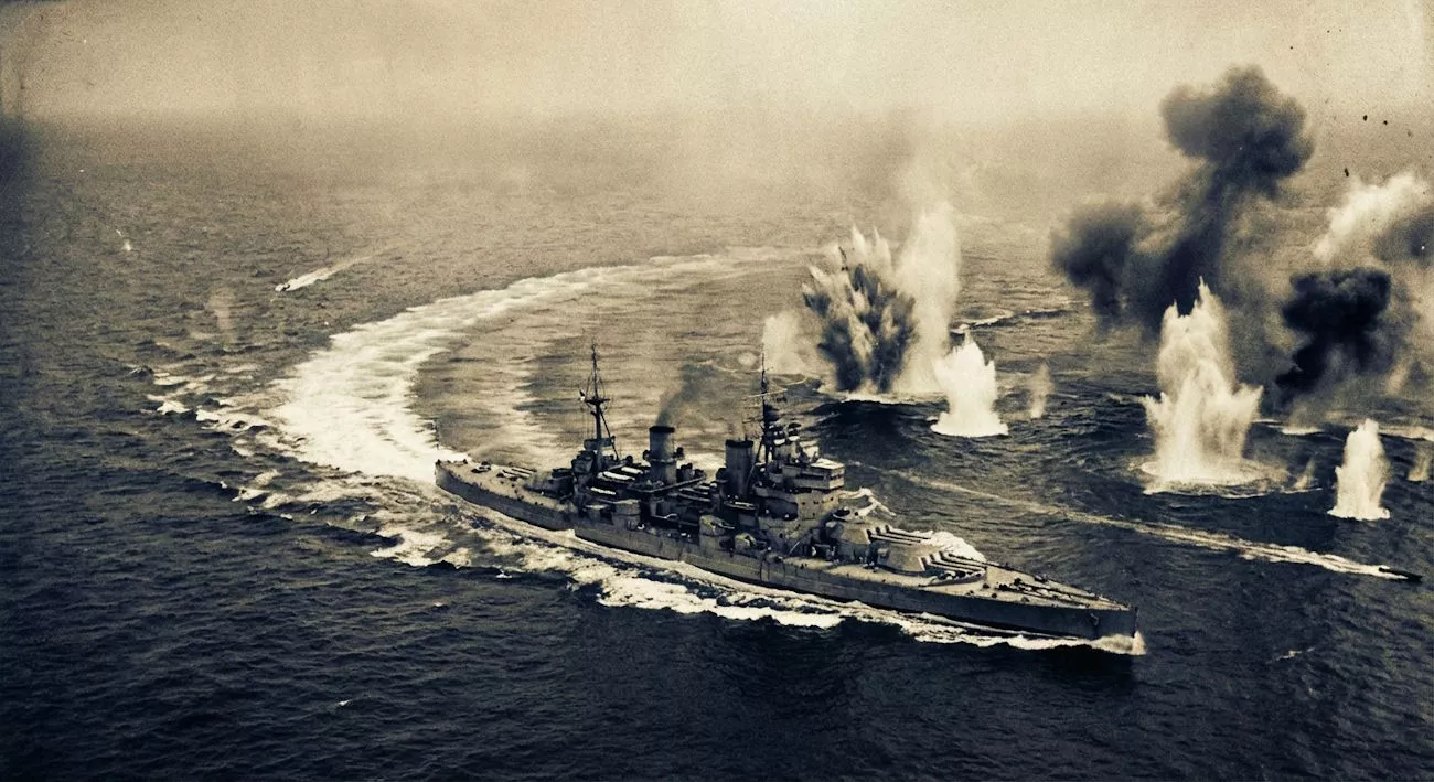 Okręt wojenny manewruje po wzburzonym morzu, otoczony eksplozjami i wodą tryskającą w powietrze. Scena odbywa się na otwartym oceanie, w czasach wojennych.
