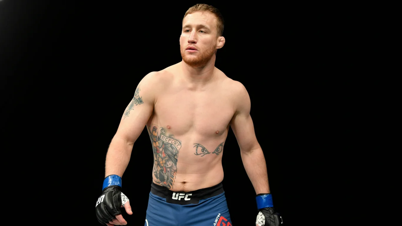 Mężczyzna z tatuażami stoi w dynamicznej pozie. Nosi niebieskie spodenki sportowe z logo UFC. Tło jest czarne.