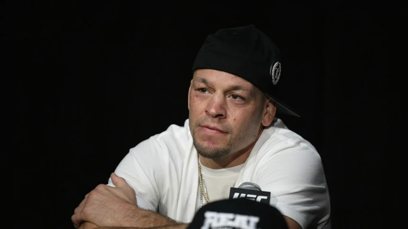 Mężczyzna w białej koszulce i czapce z daszkiem siedzi przy mikrofonie z logo „UFC” na tle czarnej kurtyny. Wygląda na zamyślonego lub skoncentrowanego.