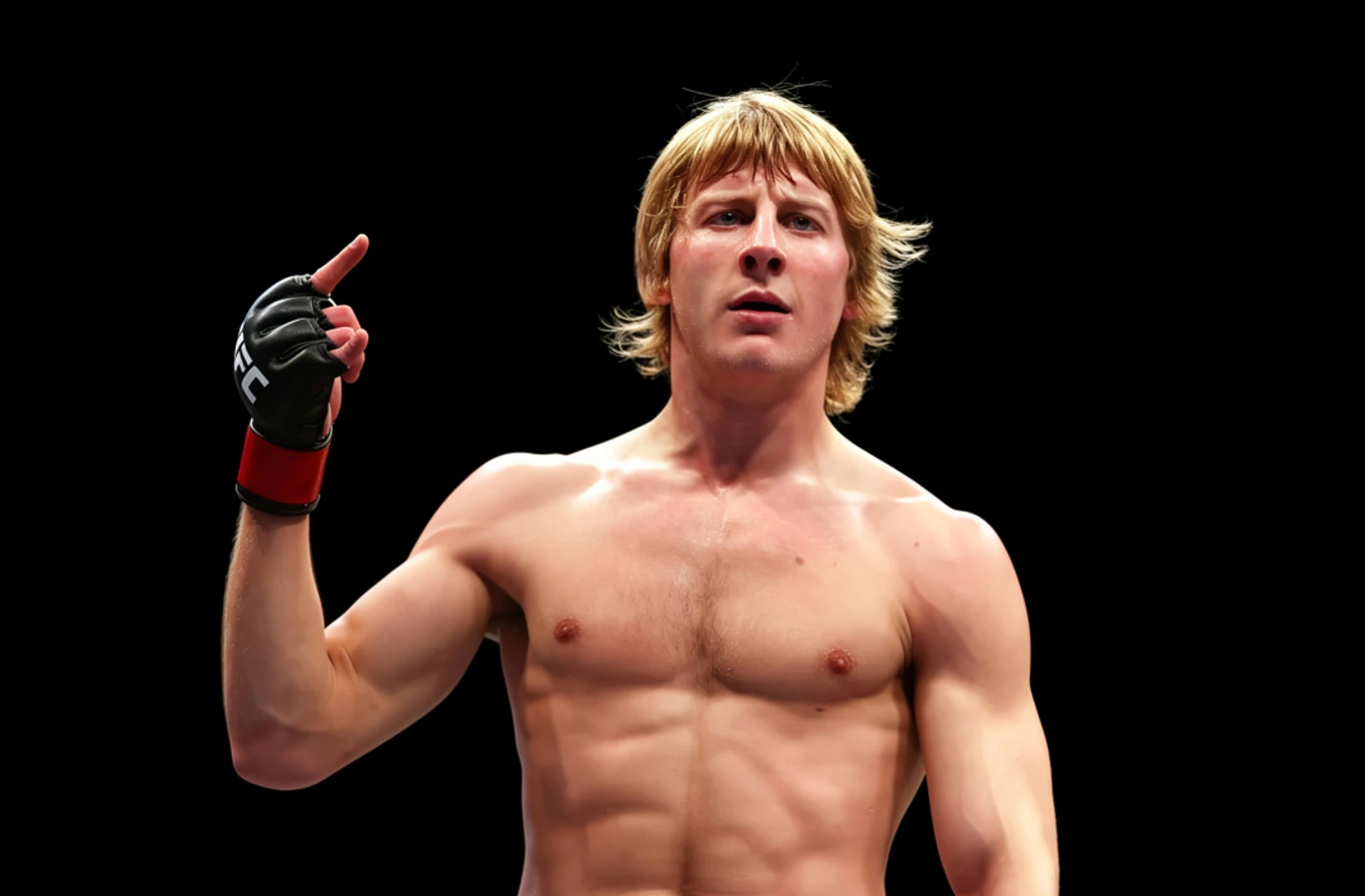 Zawodnik w rękawiczkach podnosi palec wskazujący do góry. Stoi na ciemnym tle, ukazując umięśnioną sylwetkę. Na rękawiczce widnieje napis "UFC".