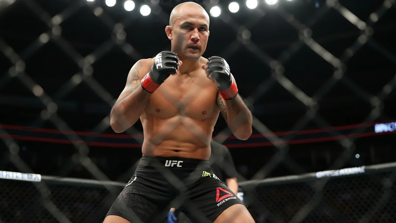 Zawodnik UFC w czarnych spodenkach stoi w pozycji obronnej w oktagonie, otoczony siatką. Światła padają z góry na arenie.