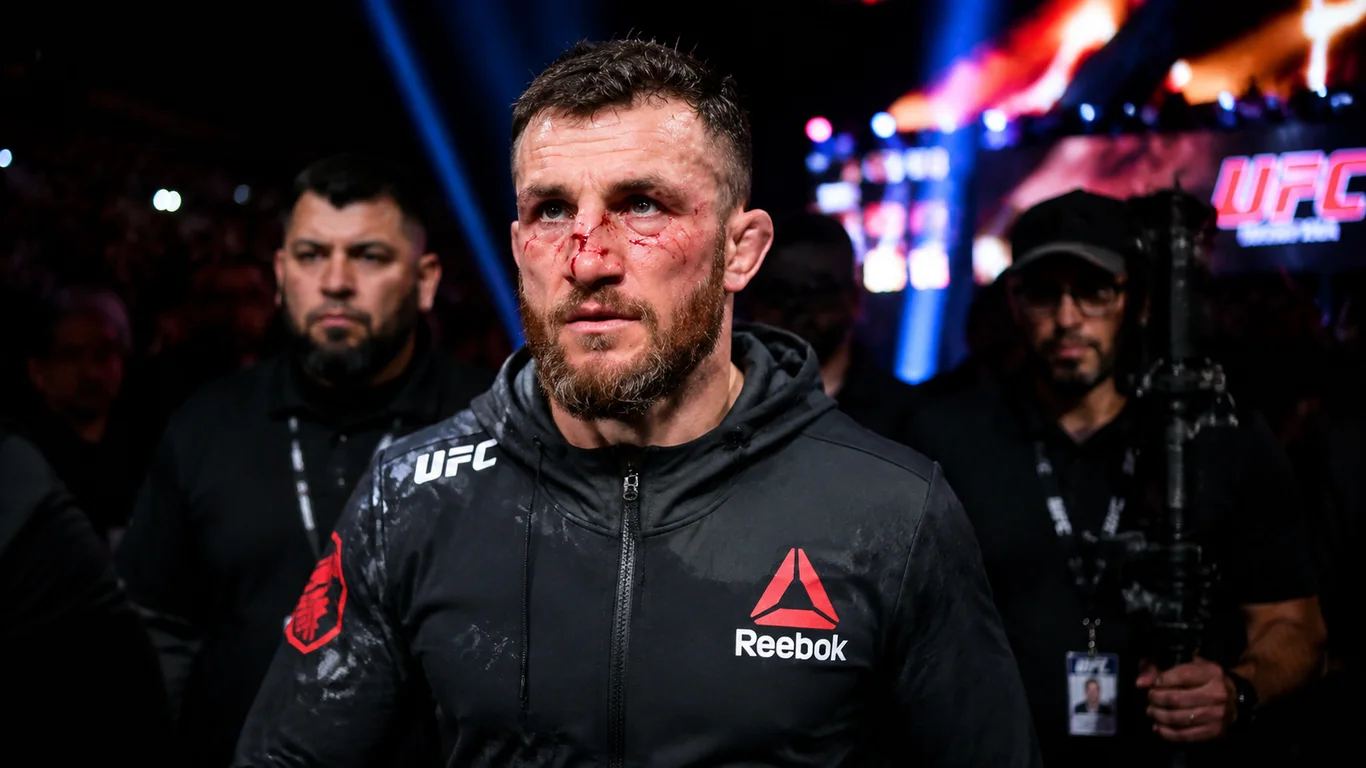 Merab Dvalishvili w hoodie UFC opuszczający oktagon po walce z krwią na twarzy, eskorta zespołu w tle gali UFC.