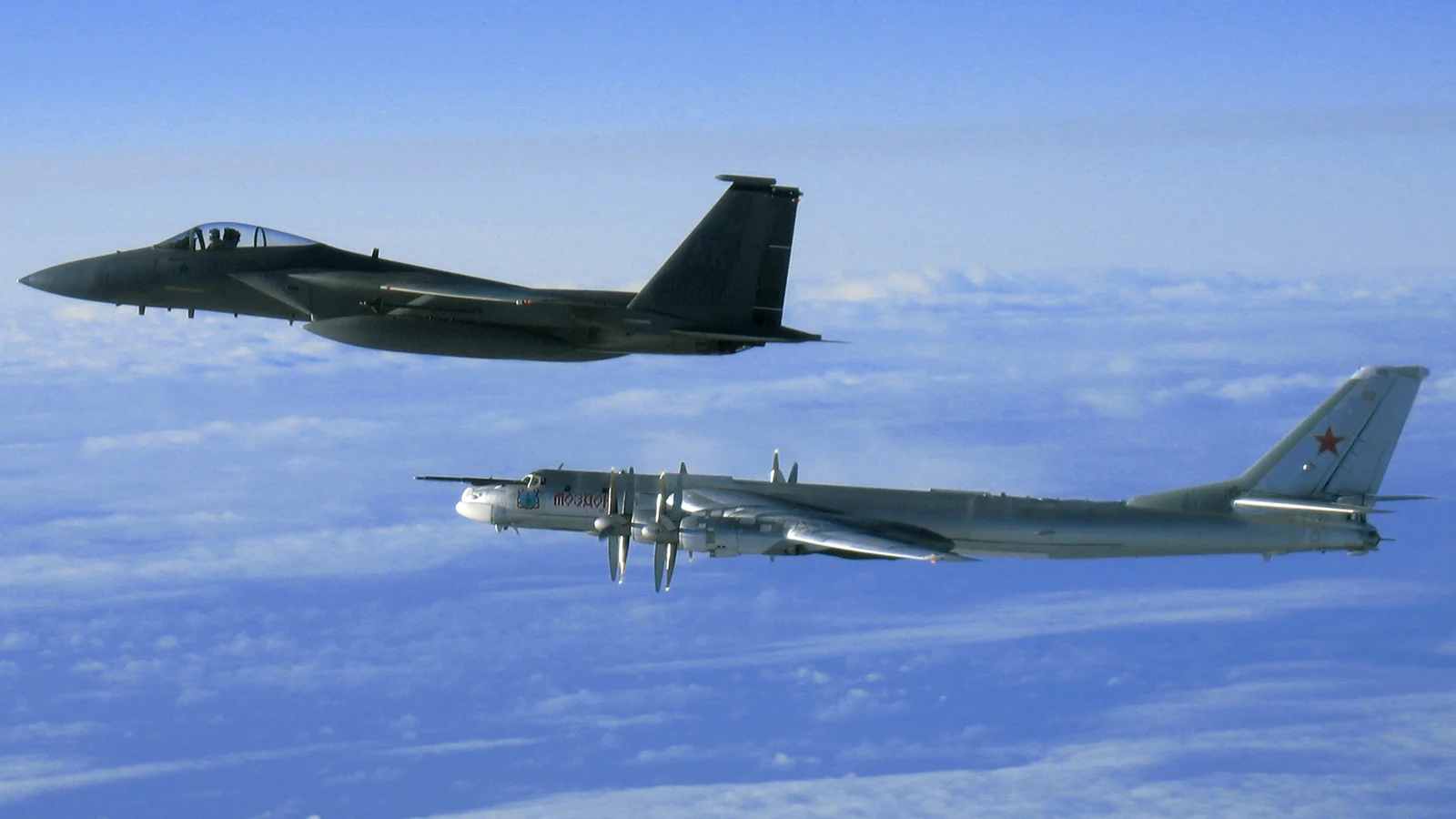 Rosyjski bombowiec Tu-95MS z napisem Mozdok na kadłubie w locie nad chmurami, obok amerykański myśliwiec F-15C w eskorcie