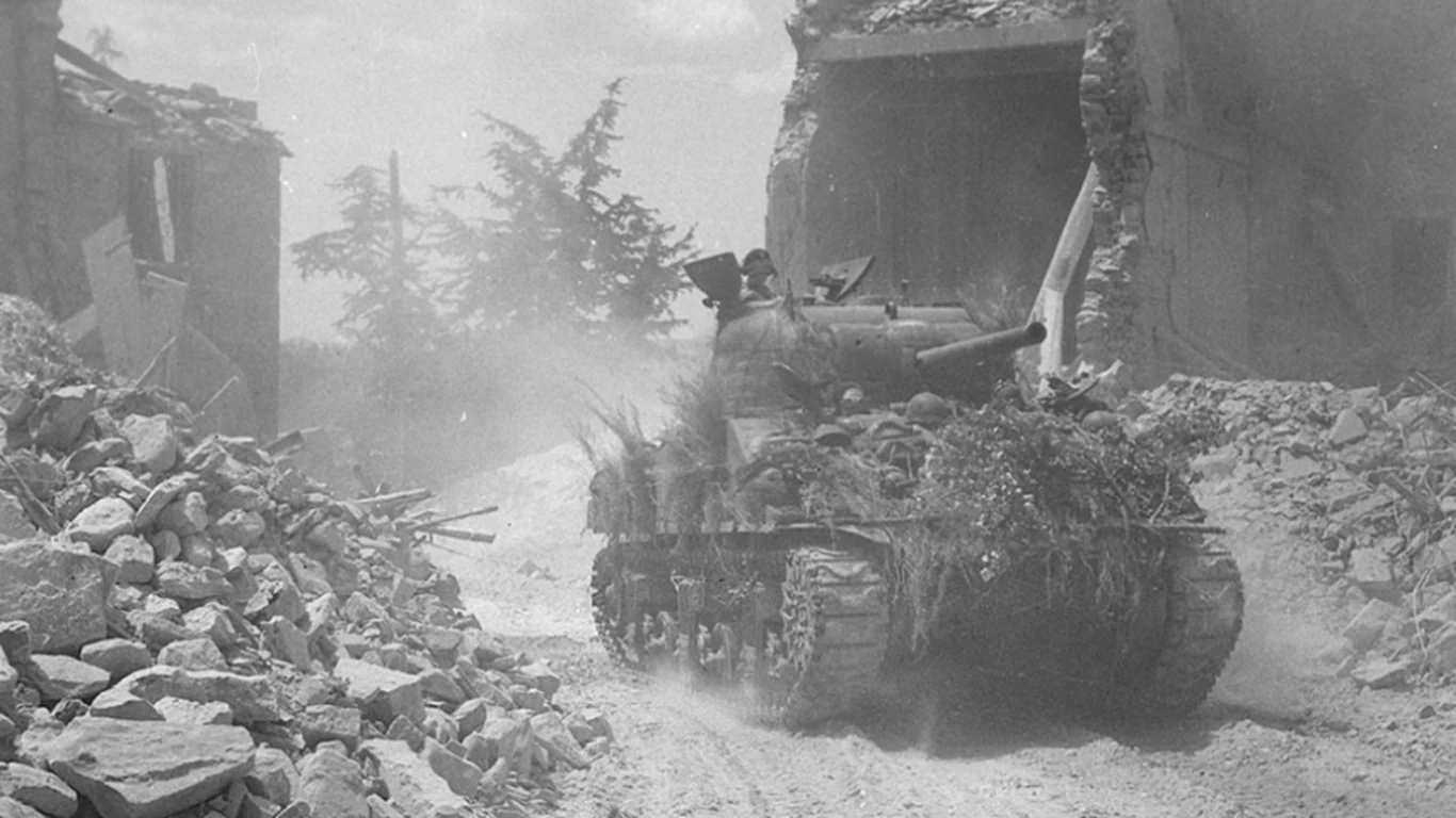 Czołg Sherman obładowany maskowaniem wjeżdża w kurzu między ruiny zabudowań włoskiego miasteczka