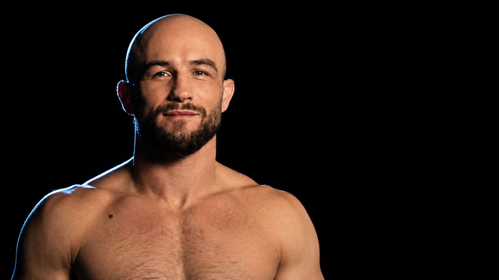 Portret zawodnika MMA z ogoloną głową i ciemnym zarostem, bez koszulki, oświetlony bocznym światłem na czarnym tle