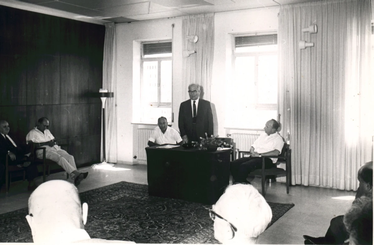 Czarno-białe zdjęcie z 1968 roku: mężczyzna w garniturze przemawia stojąc przy biurku w gabinecie, wokół siedzi kilkunastu uczestników uroczystości