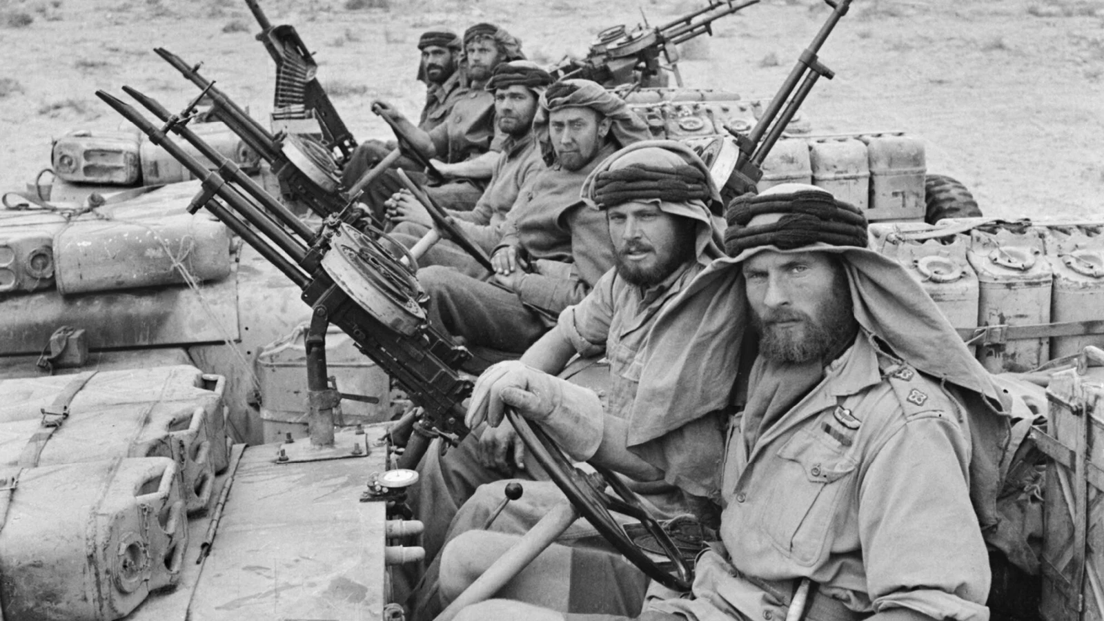 Żołnierze z brodami i w arabskich chustach siedzą w jeepach uzbrojonych w karabiny maszynowe, obwieszonych kanistrami, na tle pustyni