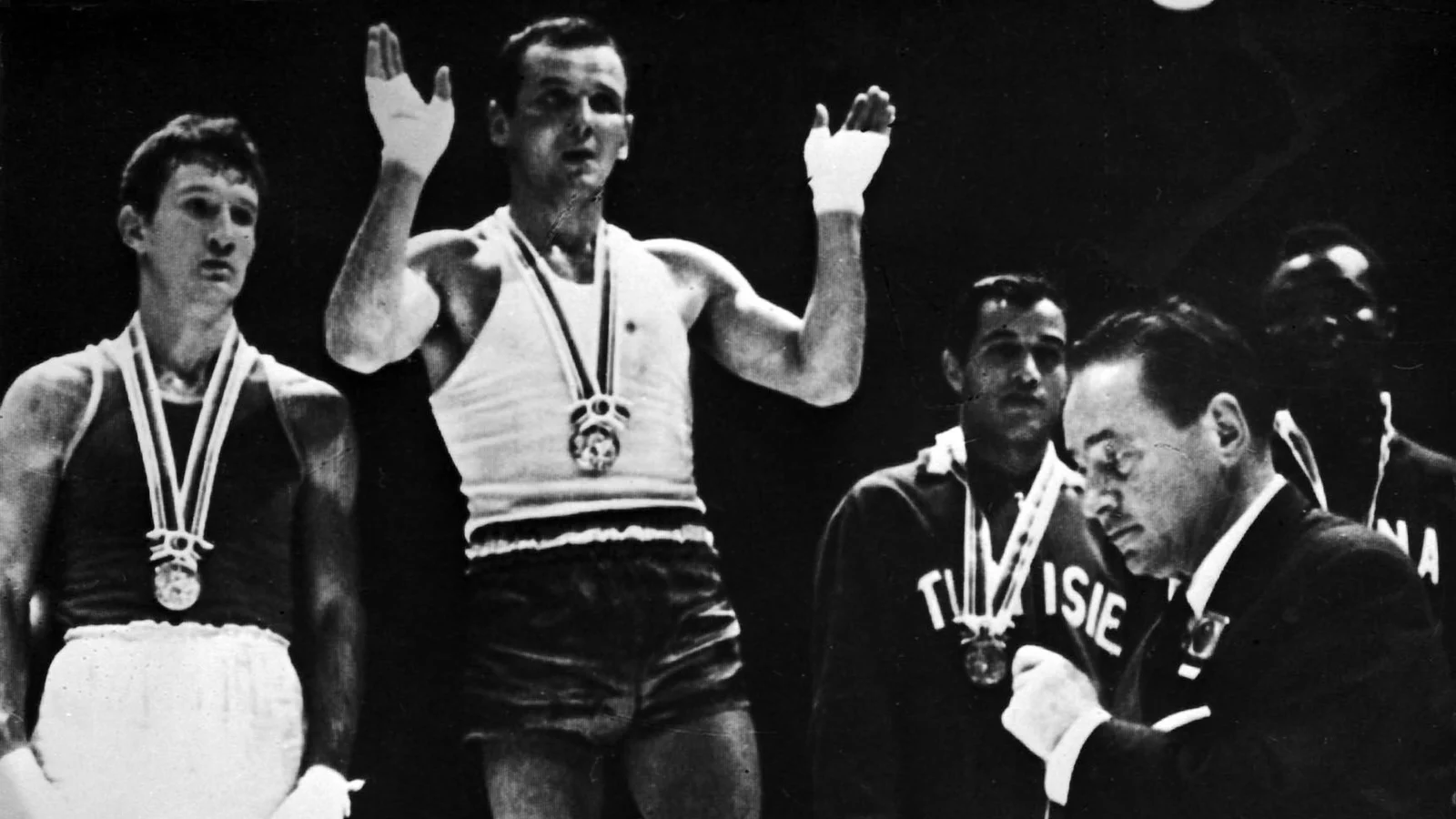Jerzy Kulej z uniesionymi rękami na najwyższym stopniu podium olimpijskiego w Tokio w 1964 roku, obok medaliści wagi lekkopółśredniej i działacz wręczający medale