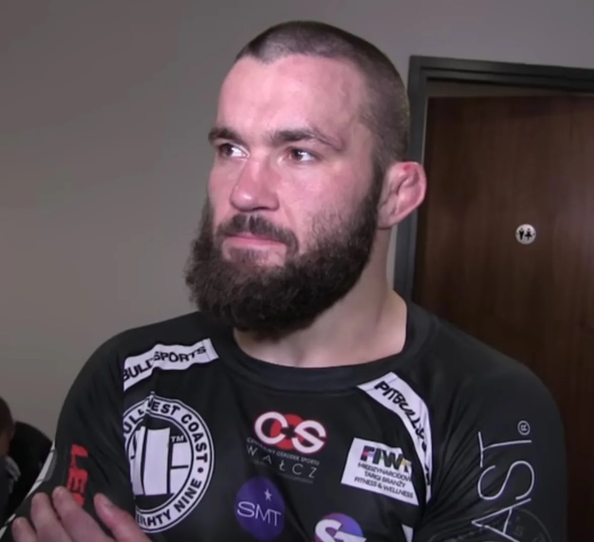 Brodaty Michał Materla w czarnej koszulce sponsorskiej, ze skrzyżowanymi rękami, podczas wywiadu na zapleczu gali