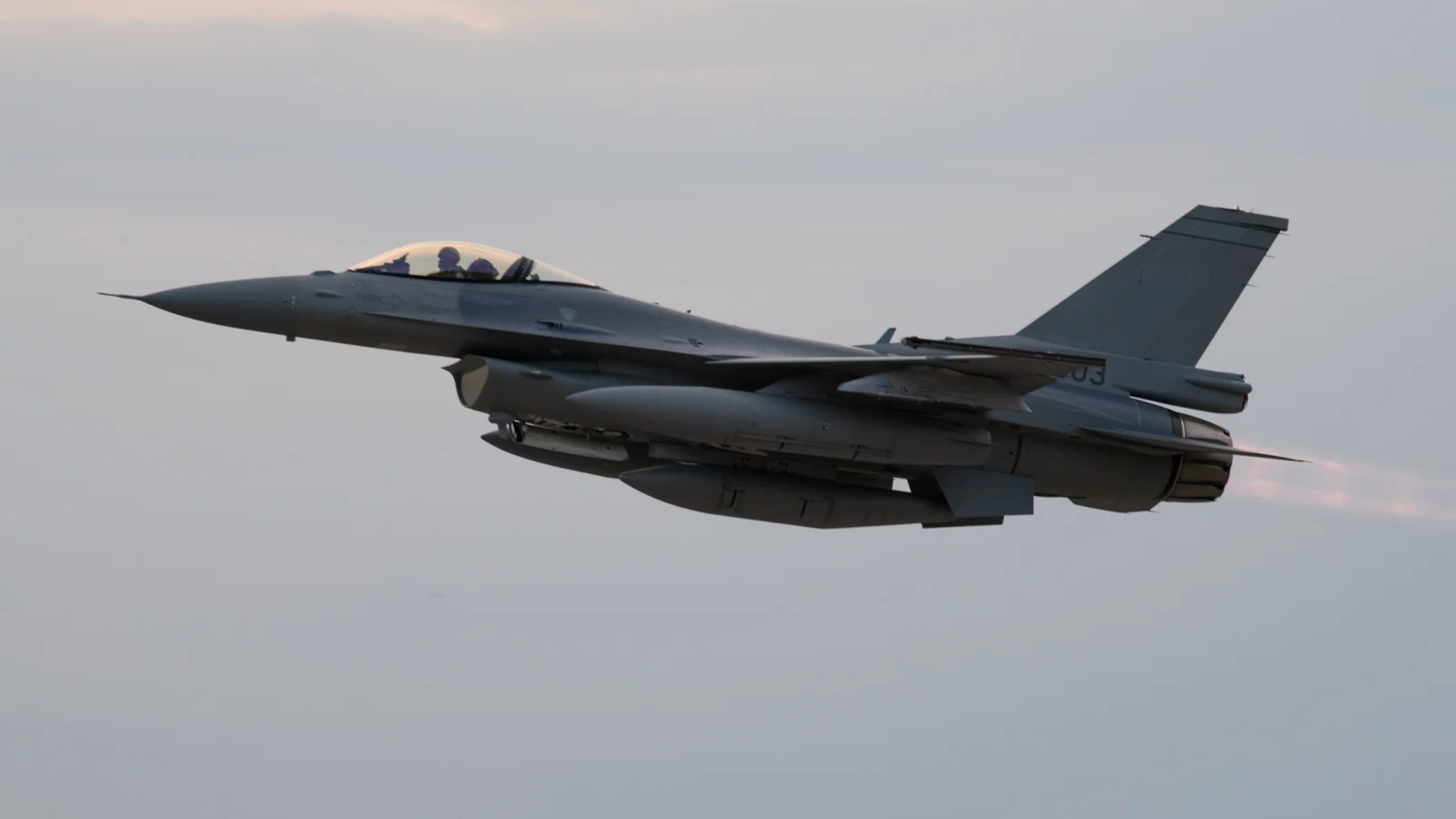 Dwumiejscowy F-16 Block 70 w locie z włączonym dopalaczem, widziany z boku na tle szarego nieba