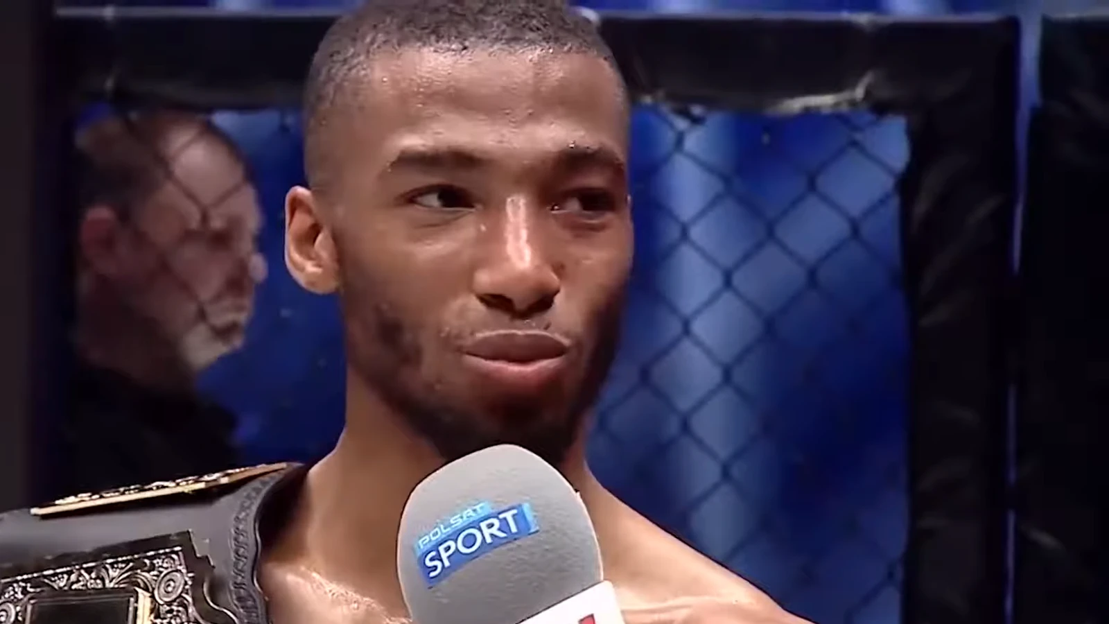 Salahdine Parnasse w klatce z mistrzowskim pasem KSW na ramieniu, przed mikrofonem Polsatu Sport