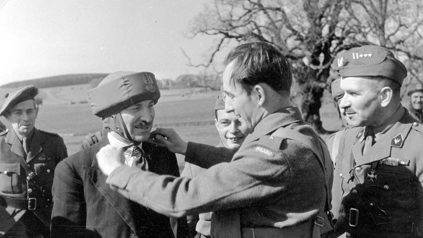 Żołnierz 1. Samodzielnej Brygady Spadochronowej zakłada Clementowi Attleemu ćwiczebny hełm spadochronowy, obok stoi uśmiechnięty płk Stanisław Sosabowski w berecie