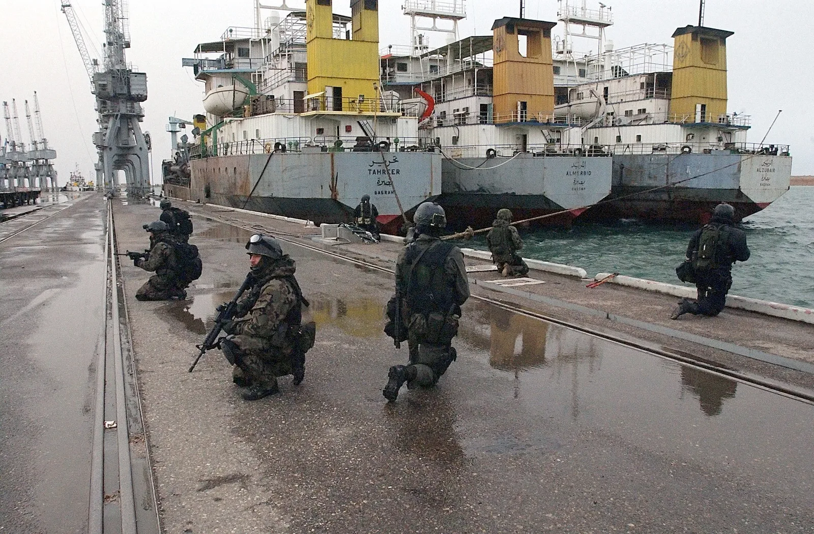 Operatorzy w hełmach i kamuflażu klęczą z karabinkami na mokrym nabrzeżu portu, za nimi zacumowane irackie pogłębiarki i dźwigi portowe