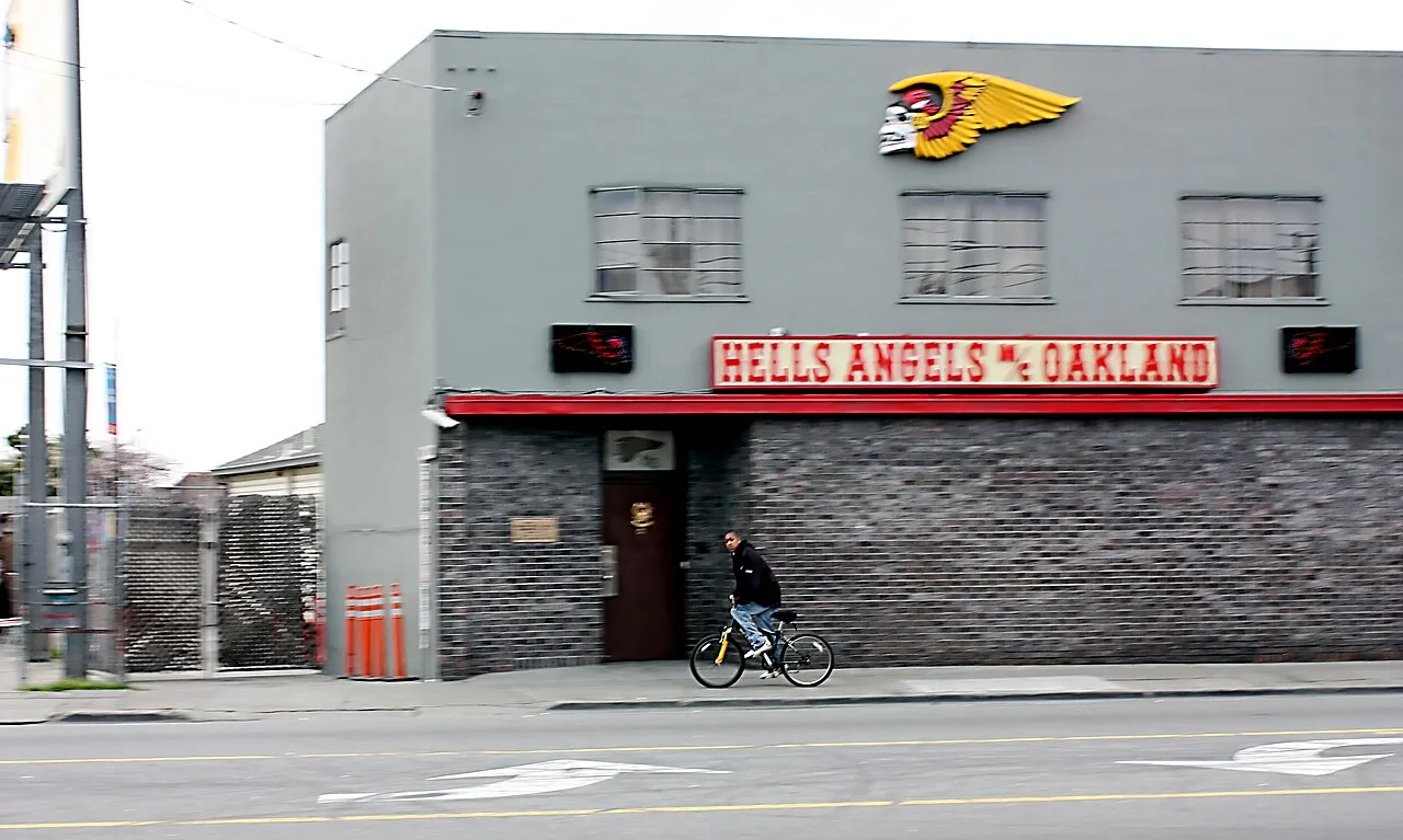 Szary piętrowy budynek z czerwonym szyldem Hells Angels MC Oakland i logo skrzydlatej czaszki na fasadzie, przed nim ulica i rowerzysta