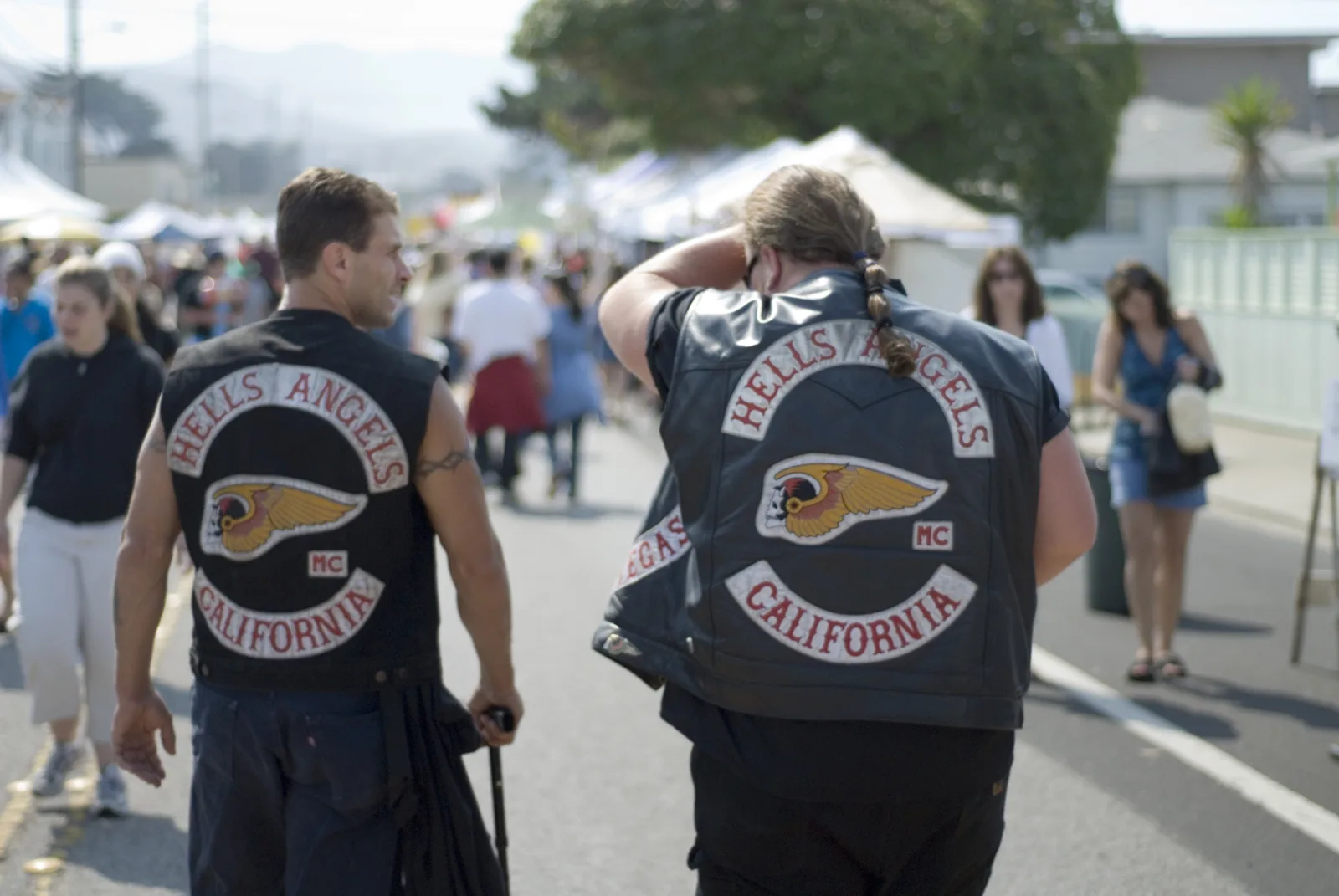 Dwóch mężczyzn w skórzanych kamizelkach z naszywkami Hells Angels California i skrzydlatą czaszką idzie tyłem do obiektywu przez zatłoczoną ulicę