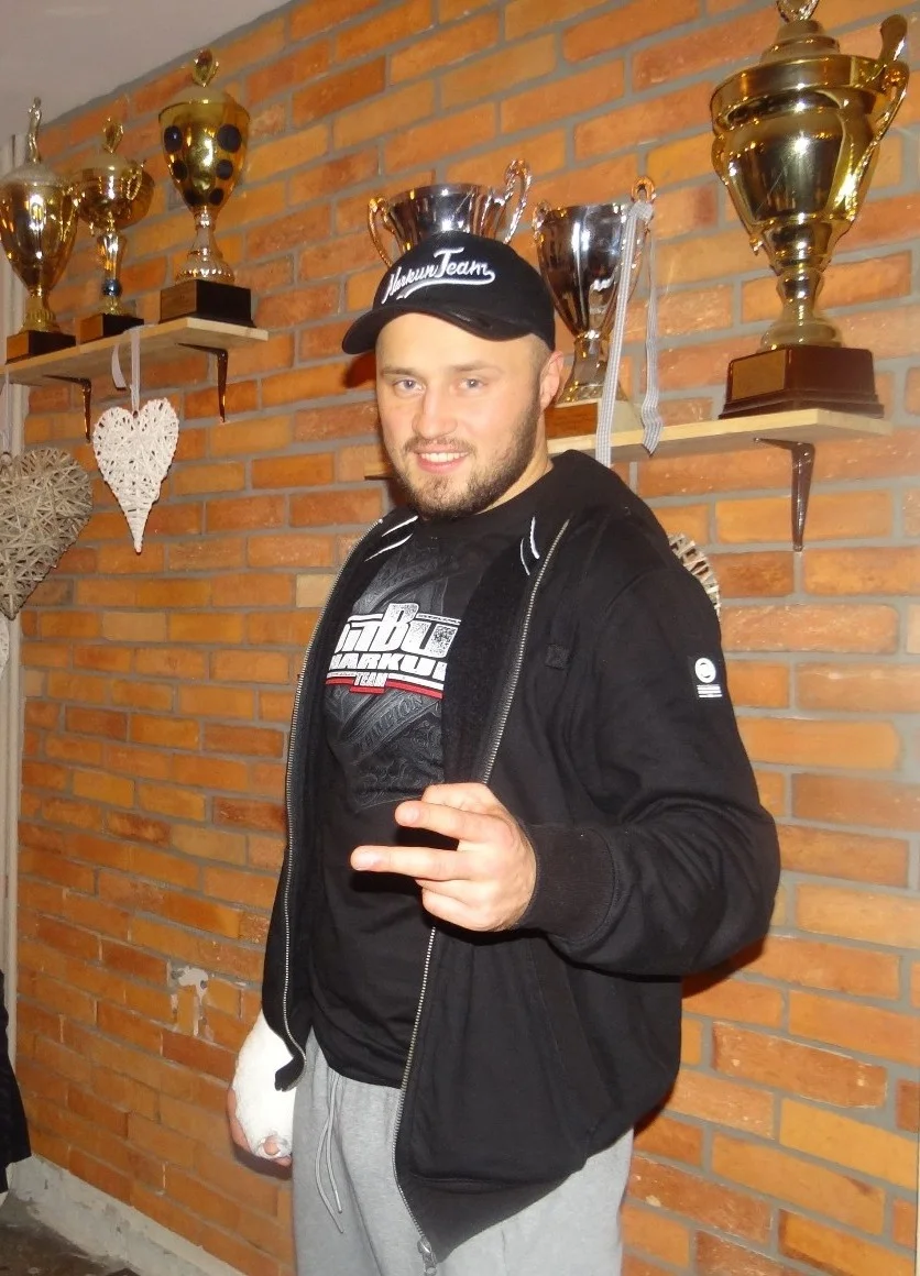 Tomasz Narkun w czapce z daszkiem i czarnej bluzie, z zabandażowaną dłonią, na tle ceglanej ściany i półek z pucharami