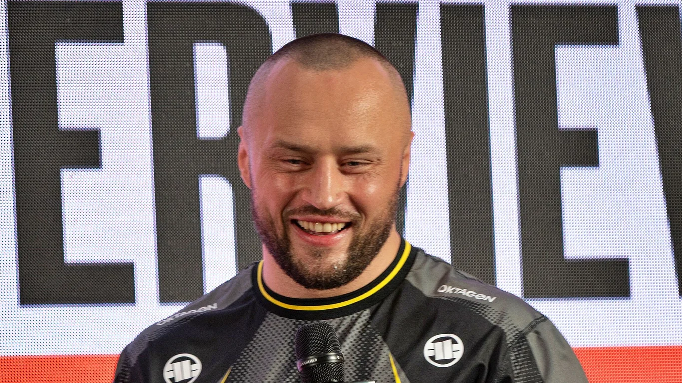 Uśmiechnięty Tomasz Narkun w czarnej koszulce Oktagonu z mikrofonem podczas ceremonii ważenia w Szczecinie