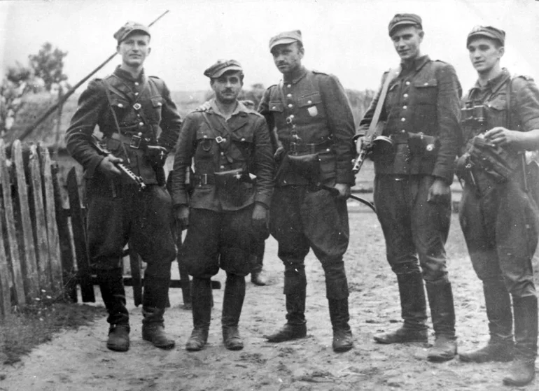 Czarno-białe zdjęcie pięciu oficerów w mundurach i rogatywkach z bronią, stojących na wiejskiej drodze przy drewnianym płocie